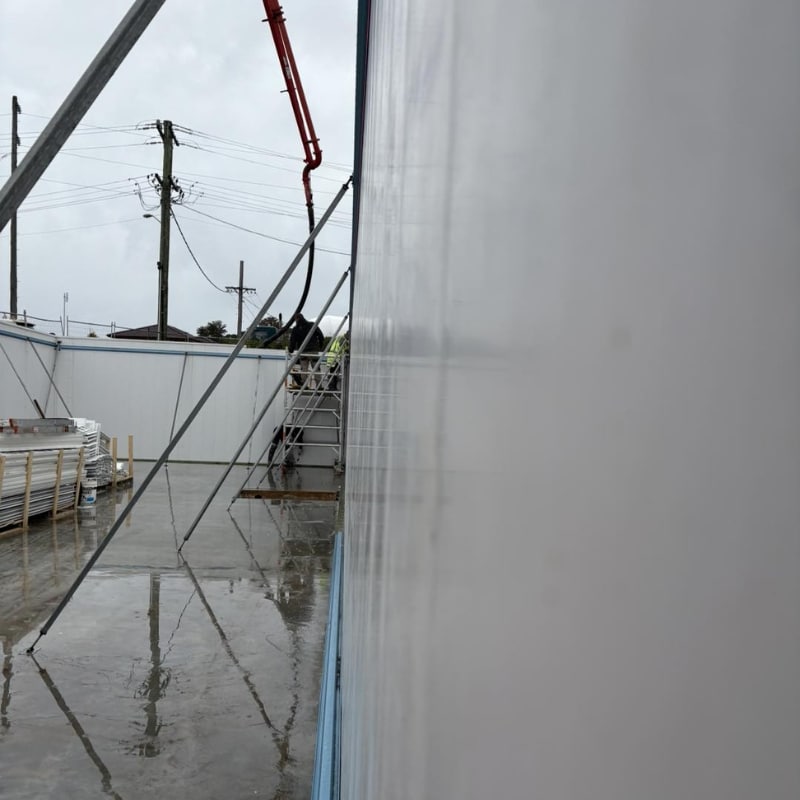 Concrete being poured into permanent formwork system with pump hose beside braced wall panels on wet construction site