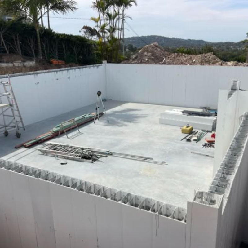 A construction site showing a white hollow permanent formwork system assembled into walls on a concrete slab foundation.